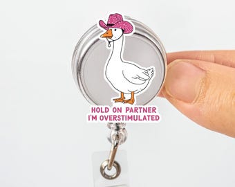 May include: A white goose wearing a pink glitter cowboy hat badge reel. The badge reel has the text "HOLD ON PARTNER I'M OVERSTIMULATED" in pink and white. The reel is silver and has a clear plastic clip.