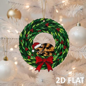 May include: A clear, 2D flat Christmas ornament. It features a sleeping calico cat wearing a Santa hat, inside a green wreath with red berries and a red bow. The ornament hangs from a gold string.