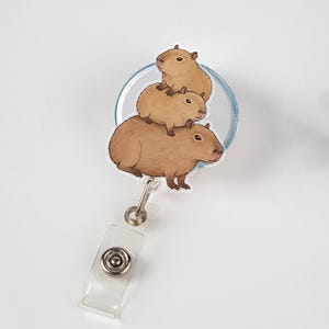 May include: A badge reel featuring a stack of three capybaras in shades of brown. The capybaras are illustrated with detailed fur and facial features. The reel has a clear plastic strap and a metal clip for attaching to clothing or accessories.