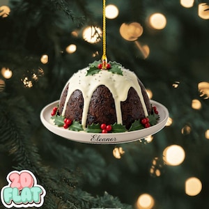 May include: A Christmas pudding ornament with white icing, holly, and red berries. The ornament is on a white plate with the name "Eleanor" and is suspended by a gold string. The background is a blurred Christmas tree.