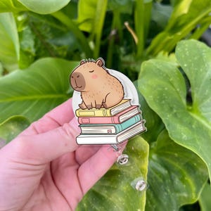 May include: A badge reel featuring a cartoon capybara sitting on a stack of books. The capybara is light brown with a dark brown nose and closed eyes. The books are yellow, pink, and blue. The reel has a clear plastic retractable cord.