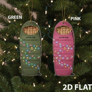 May include: Two 2D flat Christmas ornaments shaped like sleeping bags. One is green, the other pink. Both have a plaid interior, a string of colorful lights, and the text "CUSTOM NAME". The ornaments hang from gold strings.