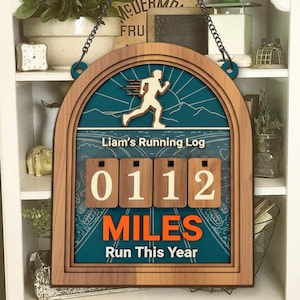 Op de afbeelding: Houten hardlooplogbord met de tekst "Liam's Running Log" en "0112 MILES Run This Year". Het bord toont een hardlopersilhouet en een teal en bruin kleurenschema. Het is ontworpen om de hardloopprogressie bij te houden.