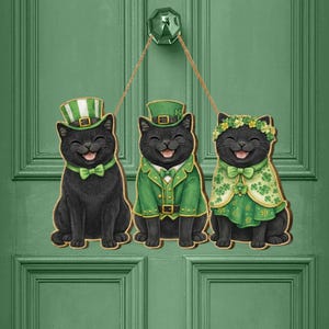 May include: A wooden St. Patrick's Day door hanger featuring three smiling black cats. Each cat is dressed in festive green attire, including hats, bow ties, and a cape, with a cheerful expression. The door hanger is suspended by a natural twine.