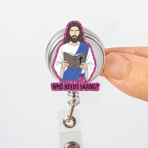 May include: A retractable badge reel featuring an illustration of Jesus holding a book with a cross. The image is outlined with pink glitter and includes the text "WHO NEEDS SAVING?" The reel has a silver metal casing and a clear plastic clip.