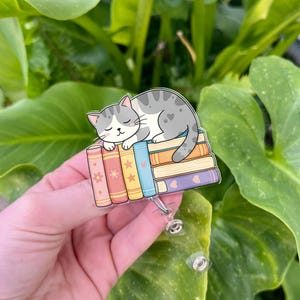 May include: A badge reel featuring a gray and white cat sleeping on a stack of colorful books. The books have floral, star, and heart designs. The reel has a clear plastic retractable cord and a metal clip.