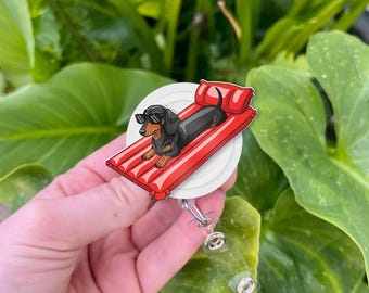 May include: A badge reel featuring a cartoon dachshund wearing sunglasses, lounging on a red pool float. The dog is black and brown, and the float has white stripes. The badge reel is white with a clear plastic retractable cord.