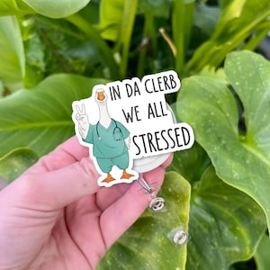 Puede incluir: Un portatarjetas blanco con un dibujo de un ganso de dibujos animados con uniforme médico turquesa y un estetoscopio. El ganso hace el signo de la paz. El texto en la tarjeta dice "IN DA CLERB WE ALL STRESSED".