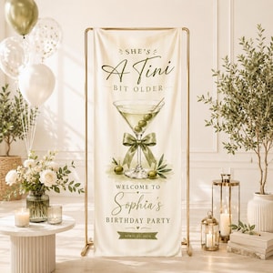 May include: A vertical cream-colored banner on a gold metal stand featuring a martini glass illustration with green olives and a green ribbon. Text reads: SHE'S A Tini BIT OLDER, WELCOME TO Sophia's BIRTHDAY PARTY, and APRIL 25, 2024.