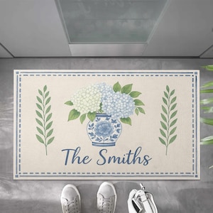 May include: A rectangular doormat with a cream background and a blue border. The mat features a blue and white floral vase with hydrangeas and the text "The Smiths". Green leaf accents are on either side of the vase.
