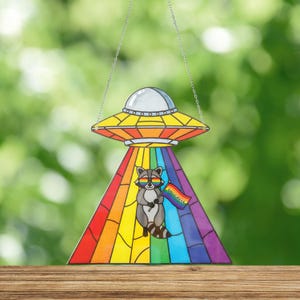 May include: A stained glass suncatcher depicting a UFO with a rainbow beam and a raccoon wearing rainbow sunglasses holding a flag with the word "PROGRESS". It hangs from a silver chain. The background is a blurred green.