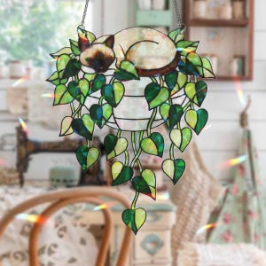 Op de afbeelding: Een gebrandschilderde kattenvanger. De kat is een Siamees ras, vredig slapend in een hangende plantenbak met groene hartvormige bladeren. De plantenbak wordt opgehangen door een zilveren ketting. De zonnevanger is ontworpen om het licht op te vangen.