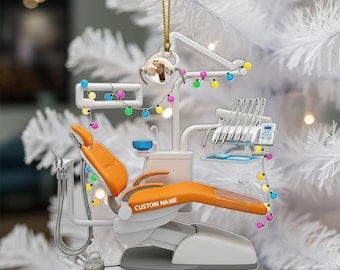 May include: A dentist's office Christmas ornament. The ornament features a miniature dental chair in orange and white, a dental light, and other dental equipment. The ornament is decorated with a string of colourful lights and has the text "CUSTOM NAME" on the chair.