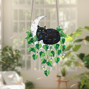 Puede incluir: Un atrapasoles de vidriera con un gato negro sobre una luna creciente, rodeado de hojas verdes y flores blancas. Cuelga de una cadena plateada y mide aproximadamente 20 cm de diámetro.
