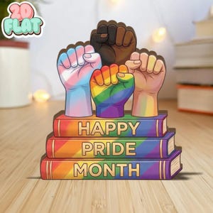 May include: A colorful Pride Month decoration featuring raised fists in various skin tones and a rainbow-colored fist. The fists are atop three books with the words "HAPPY PRIDE MONTH" in gold lettering. The image includes a "2D FLAT" sticker.