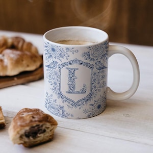 May include: White ceramic mug with a blue floral and bird design. The mug features the letter "L" in a decorative shield. The mug is filled with a warm beverage and is next to pastries.