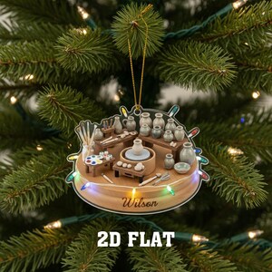 May include: A circular Christmas tree ornament depicting a pottery studio. The ornament shows miniature pottery wheels, shelves with ceramic pots, and a paint palette. The name "Wilson" is printed on the wooden base, with the text "2D FLAT" below.