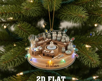 May include: A round, clear acrylic Christmas ornament featuring a pottery studio scene. The ornament includes miniature pottery wheels, shelves with ceramic pots, and a paint palette. The name "Wilson" is printed on the wooden base, with the text "2D FLAT" below.
