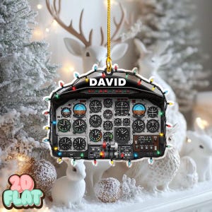 May include: A Christmas ornament shaped like an airplane cockpit, featuring numerous gauges and dials. The name "DAVID" is displayed at the top. The ornament is decorated with colorful Christmas lights and is hanging in front of a winter scene.