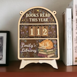 May include: Wooden bookshelf decor with the text "BOOKS READ THIS YEAR" and "Emily's Library." The decor features a cat sleeping on books and the number "112." The background has a floral pattern.