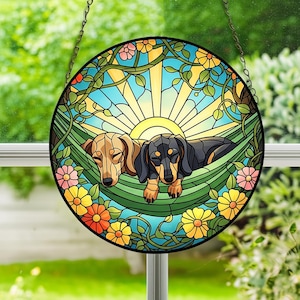Könnte beinhalten: Rundes Buntglasfenster mit zwei Dackeln, die sich in einer Hängematte entspannen. Das Kunstwerk hat eine lebendige Farbpalette mit blauem Himmel, gelber Sonne und bunten Blumen. Das Stück ist mit einem schwarzen Rahmen versehen und hat eine Aufhängekette.