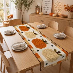 May include: A rectangular fabric table runner featuring a repeating pattern of orange and cream-colored pumpkins with green stems and small black stars on a white background, bordered by a checkered orange and brown fabric trim on a wooden table.