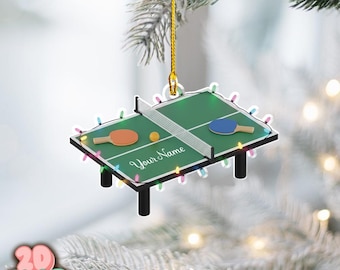 May include: A green ping pong table Christmas ornament with a white net, two paddles, and a ball. The ornament has the text "Your Name" and is decorated with colourful lights. The ornament is hanging from a gold string.