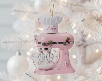 May include: A pink kitchen mixer Christmas ornament with the name "Lily" on a chef's hat. The ornament has a clear bowl with whisks and is decorated with floral accents and star details. The ornament is hanging on a white Christmas tree.