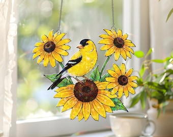 May include: A decorative window hanging featuring a yellow bird perched on a sunflower. The bird has a black head and wing markings. The sunflowers are yellow with brown centers and green leaves. The hanging is suspended by a silver chain.