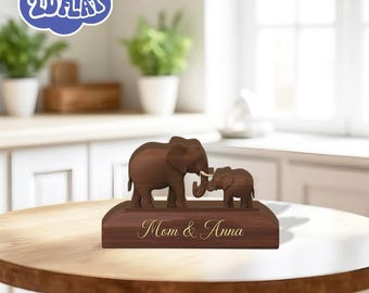 May include: A wooden sculpture of two elephants, a larger one and a smaller one, touching trunks. The base is engraved with "Mom & Anna" in gold lettering. The sculpture is set on a wooden table.