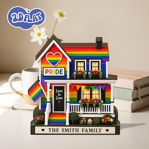 Puede incluir: Figurita de casa Pride colorida con techo arcoíris, logotipo en forma de corazón y la palabra "PRIDE". La casa tiene un porche con una pancarta que dice "Love Is Love". La base dice "THE SMITH FAMILY".