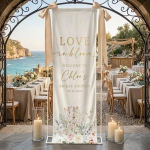 May include: A cream-colored banner with gold text that reads "LOVE in bloom" and "Welcome to Chloe's Bridal Shower 08.20.2026." The banner is decorated with floral arrangements and tied with beige ribbons. The scene is set outdoors with tables and ocean views.