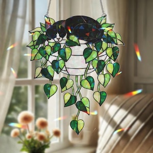 Pode incluir: Um apanhador de sol decorativo pendurado com um gato e uma planta. O gato é preto e branco, descansando sobre um vaso com folhas verdes em forma de coração. O apanhador de sol é pendurado por uma corrente prateada.
