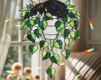 May include: A decorative hanging cat and plant suncatcher. The cat is black and white, resting on a pot of green heart-shaped leaves. The suncatcher hangs from a silver chain and is displayed in front of a window.