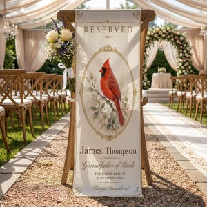May include: A vertical cream-colored fabric sign draped over a wooden chair, featuring the text RESERVED, James Thompson, Grandfather of Bride, Your love still guides us as we celebrate today, Always Remembered, and a red cardinal bird illustration.