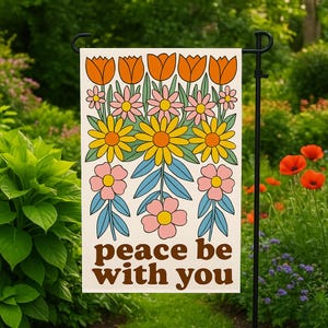 Op de afbeelding: Een tuinvaandel met een retro bloemenontwerp in oranje, geel, roze en blauw. De vlag toont de woorden "peace be with you" in bruine letters. De vlag staat in een tuin.