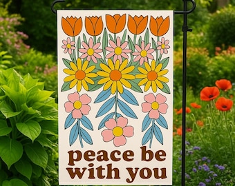 May include: A garden flag with a floral design in orange, yellow, pink, and blue. The flag has the words "peace be with you" in brown lettering. The flag is displayed in a garden setting.