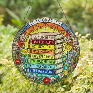 May include: Circular stained-glass style hanging decoration with the text "IT IS OKAY TO" at the top. Below are stacked books with phrases like "BE YOURSELF" and "START OVER AGAIN." The design includes floral and sunburst elements.