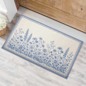 May include: Rectangular doormat with a cream-colored background and a blue floral design. The border and floral details are in a soft blue hue. The mat is placed on a light-colored wooden floor near a wooden door.