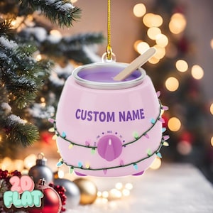 Może przedstawiać: Różowa ozdoba choinkowa w kształcie podgrzewacza do wosku, z fioletowym napisem "CUSTOM NAME". Posiada fioletowe wnętrze, drewniany patyczek i jest ozdobiona sznurem kolorowych światełek. Pokrętło wskazuje ustawienia temperatury.