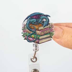 May include: A badge reel featuring a teal dragon resting on a stack of books. The dragon has blue wings with pink accents. The books are brown, purple, and green, with floral accents. The reel has a clear plastic front.