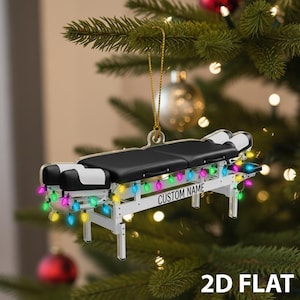 May include: A 2D flat Christmas ornament shaped like a massage table. The table is white with a black top and black and white headrests. It is decorated with colorful string lights and the text "CUSTOM NAME".
