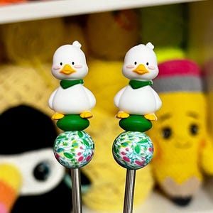 May include: Two duck-themed knitting needle stoppers. Each stopper features a white duck with a green scarf and yellow feet atop a floral patterned bead. The needles are silver in color.