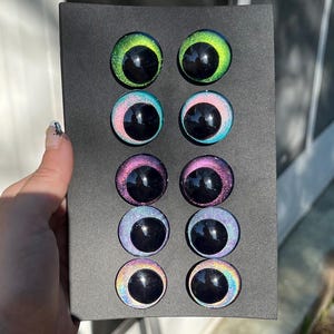 May include: A black rectangular box holding eight colorful craft eyes. The eyes have a glossy finish with a black pupil and iridescent colors including green, blue, pink, and rainbow hues. The eyes are arranged in two rows of four.