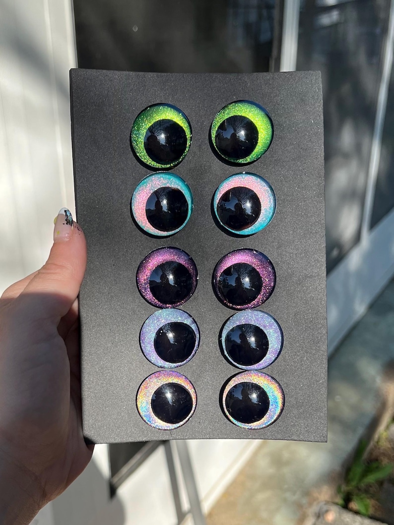 May include: A black rectangular box holding eight colorful craft eyes. The eyes have a glossy finish with a black pupil and iridescent colors including green, blue, pink, and rainbow hues. The eyes are arranged in two rows of four.