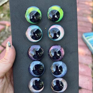 May include: A collection of ten glass eye cabochons in various colors, including green, blue, purple, and silver. The eyes are round with a reflective, glossy finish, and are displayed on a black background.