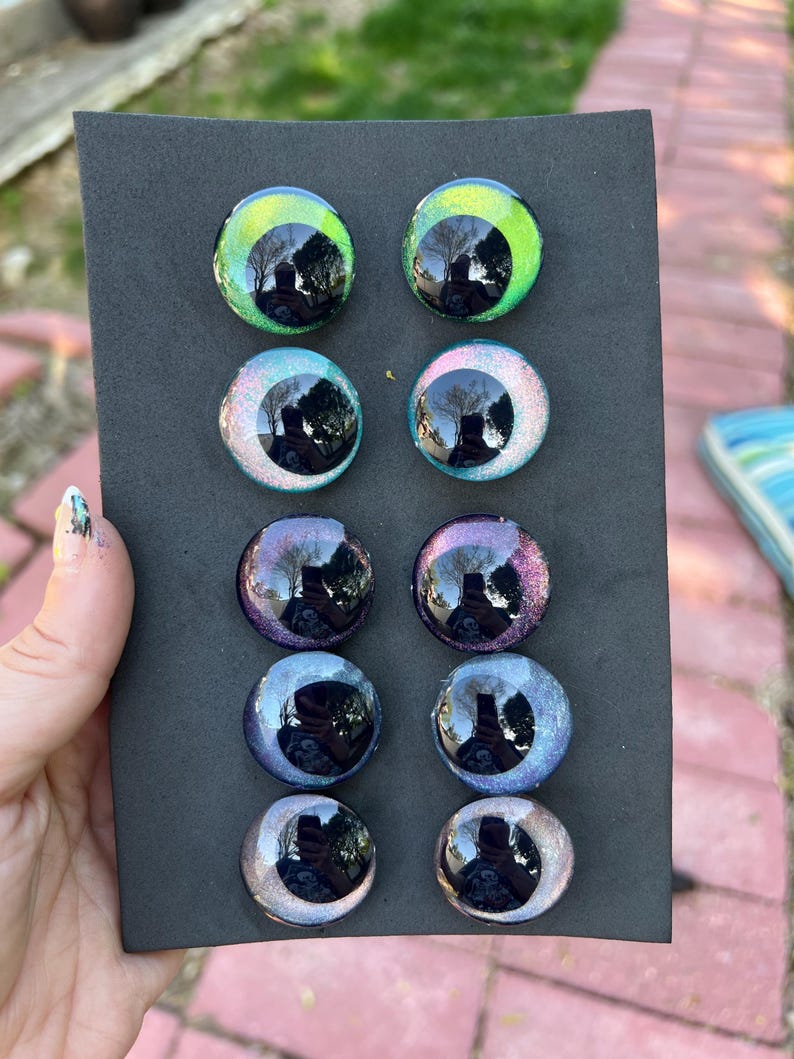 May include: A collection of ten glass eye cabochons in various colors, including green, blue, purple, and silver. The eyes are round with a reflective, glossy finish, and are displayed on a black background.