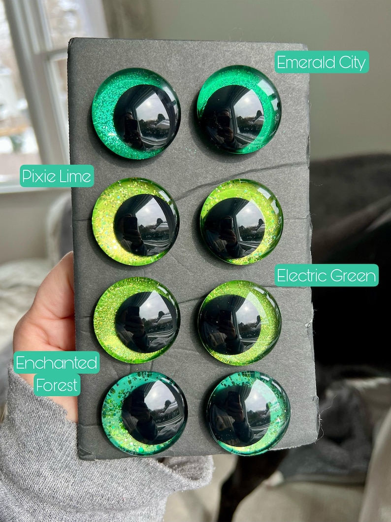 May include: A set of eight green and black glass eyes with glitter. The eyes are arranged in two rows of four. The eyes are labeled with the names "Emerald City", "Pixie Lime", "Electric Green", and "Enchanted Forest".