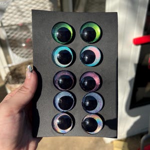 May include: A black card displays eight glass eye cabochons in various colors, including green, blue, purple, and iridescent rainbow hues. Each eye has a black pupil and a reflective, glossy finish, suitable for crafting or doll making.