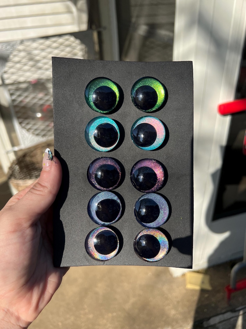 May include: A black card displays eight glass eye cabochons in various colors, including green, blue, purple, and iridescent rainbow hues. Each eye has a black pupil and a reflective, glossy finish, suitable for crafting or doll making.